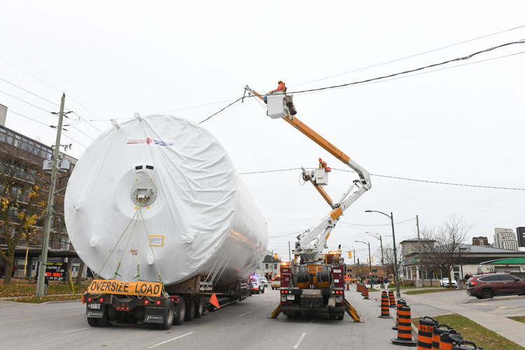 Linesperson lifting powerlines to help tank on flatbed truck being driven through the city